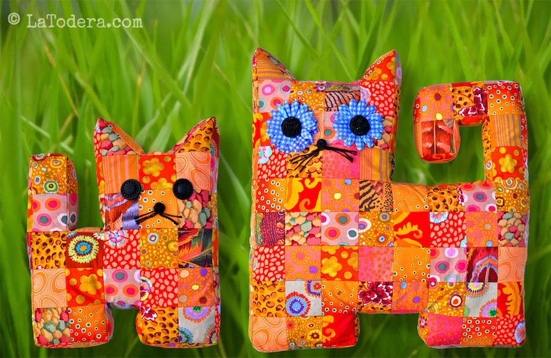 May include: Two patchwork fabric cat toys. The larger cat is orange, pink, and blue with a black nose and blue eyes. The smaller cat is orange, pink, and brown with a black nose and black eyes. Both cats are sitting on a green grassy surface.