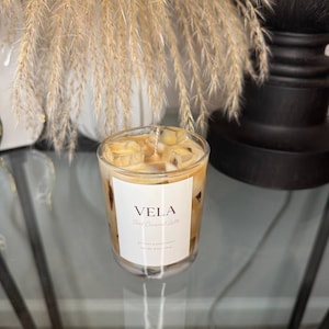 May include: A clear glass candle with a caramel-colored wax, resembling an iced caramel latte. The candle has a white label with the word "VELA" and "iced caramel latte" printed on it. The candle is on a glass surface.