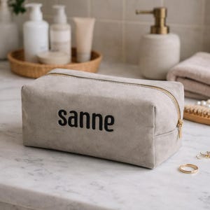 Op de afbeelding: Een rechthoekige, lichtgrijze make-uptas met de naam "sanne" in het zwart geborduurd. De tas heeft een gouden rits en staat op een marmeren oppervlak. Andere badkameraccessoires staan op de achtergrond.