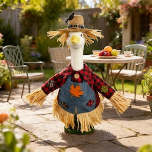 Può includere: Una statuetta decorativa a forma di oca vestita da spaventapasseri, con un cappello di paglia con un uccellino nero, una camicia a quadri rossi e neri e una salopette di jeans blu decorata con una foglia d'acero in feltro e toppe.