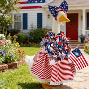 May include: A goose dressed in patriotic attire, including a red and white striped dress, a bow with stars, and a star-studded garland. It holds a small American flag, perfect for Independence Day celebrations.