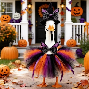 May include: A white goose wearing a black witch hat with purple glittery bat wings and a matching bow. The goose also wears a layered tulle tutu in orange, black, and purple, featuring a small bow with purple bat wings at the neck.