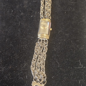 May include: A rectangular wristwatch with a silver-tone face and gold-tone accents on the top and bottom of the case. The watch features a multi-strand chain link bracelet made of silver-tone metal, with a small gold-tone crown on the right side.
