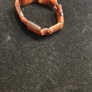 May include: A stretch bracelet composed of rectangular, polished stone beads. The beads feature banded patterns in shades of deep orange, reddish-brown, and translucent white, arranged in a circular loop to form a continuous, flexible jewelry piece.