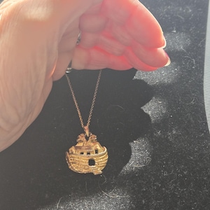 Könnte beinhalten: Eine goldfarbene Metall-Halskette mit einem Anhänger, der ein detailliertes Boot mit zwei Tierfiguren auf dem Deck zeigt. Der Anhänger hängt an einer feinen goldfarbenen Kette und weist eine strukturierte Oberfläche am Schiffsrumpf auf.