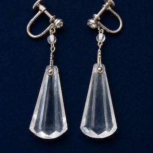 May include: A pair of silver-toned screw-back earrings featuring clear, faceted, trapezoidal glass pendants. Each earring is suspended from a small, round, clear stone set in a silver-toned metal frame, connected by a short, delicate silver-toned chain.