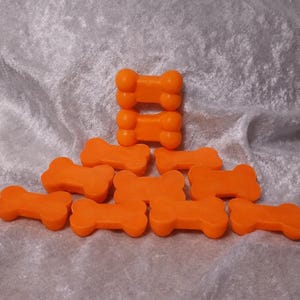 Peut inclure: Des friandises pour chiens en forme d'os, de couleur orange vif, sont disposées sur une surface texturée gris argenté. Certaines friandises sont empilées, d'autres éparpillées. Les friandises sont d'une couleur unie avec des bords arrondis.