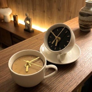 May include: Two white ceramic teacups repurposed as clocks. One cup features a black clock face with gold hands, while the other has a tan clock face with gold hands. Both cups have matching handles and are positioned on a wooden surface.