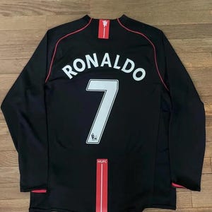 May include: Black long-sleeved football shirt with the name "RONALDO" and the number "7" in white. Red trim details the collar, sleeves, and a vertical stripe. The MUFC logo is visible.