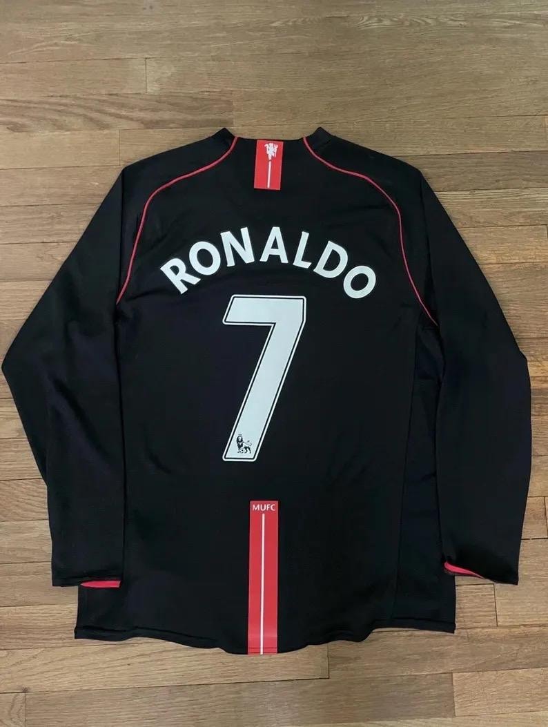 May include: Black long-sleeved football shirt with the name "RONALDO" and the number "7" in white. Red trim details the collar, sleeves, and a vertical stripe. The MUFC logo is visible.