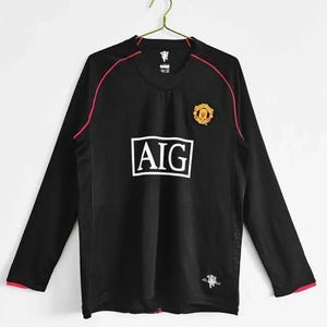 May include: Black long-sleeved football shirt with red trim and accents. The shirt displays the AIG logo in white, a team crest, and a small emblem. The shirt is hanging on a gold hanger.