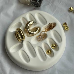 Könnte beinhalten: Goldfarbener Schmuck, darunter Creolen, Ohrstecker und eine Halskette mit Anhänger, auf einem weißen Keramiktablett präsentiert. Im Hintergrund sind zwei kleine Glasflaschen mit Korken zu sehen.