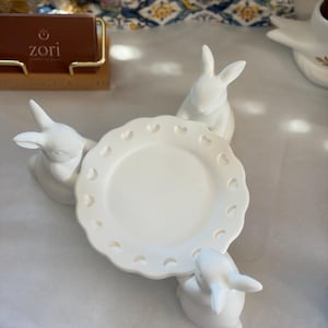 Op de afbeelding: Een witte keramische plaat met een geschulpte rand en hartvormige uitsparingen, ondersteund door drie witte konijnenbeeldjes. De plaat en konijnen staan op een wit oppervlak, met een vage achtergrond.