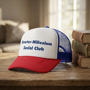 Op de afbeelding: Truckerpet met een rode klep, wit voorpaneel en blauwe mesh achterkant. De tekst "Quarter-Millennium Social Club" is in blauw op het voorpaneel gedrukt. De pet ligt op een houten oppervlak.