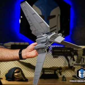 May include: A grey, 3D-printed model of a spacecraft, held in a person's hand. The model has detailed wings and a cockpit. The background includes a blue and white helmet design and the text "PATREON.COM/GALACTICARMORY".
