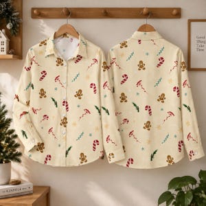 May include: Two long-sleeve button-down shirts in a light cream colour featuring a repeating holiday print of gingerbread men, red and white candy canes, green pine sprigs, and small yellow snowflakes scattered across the fabric.