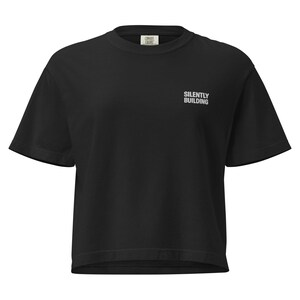 May include: Black short-sleeved t-shirt with the text "SILENTLY BUILDING" in white on the front. The shirt has a crew neck and a small label at the top. The shirt is a solid black color.