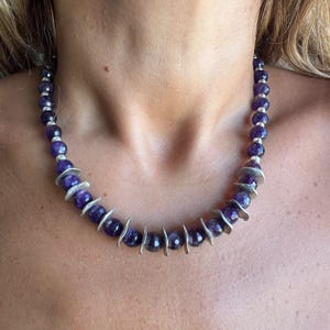 May include: A necklace featuring faceted purple amethyst beads alternating with small, curved silver-toned metal spacers. The beads are round and have a polished, textured surface, creating a consistent pattern around the entire length of the necklace.