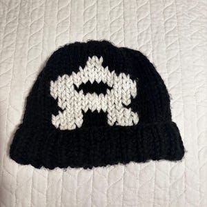 May include: A black knit beanie featuring a white, stylized graphic in the center. The hat is made of a thick, textured yarn, and the design is prominently displayed against the dark background. The beanie has a folded brim.