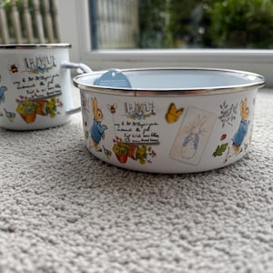 Puede incluir: Juego de taza y cuenco de esmalte blanco con ilustraciones de un conejo con chaqueta azul, flores y mariposas. El juego tiene un borde plateado e incluye texto. La taza tiene un asa.