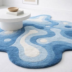 May include: A blue and white abstract rug with an organic shape. The rug has a layered design with varying shades of blue and white, creating a flowing, water-like pattern. The rug is on a white floor, with a white round coffee table and a wooden bowl on top.