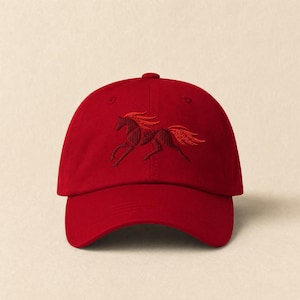 May include: A red baseball cap with an embroidered running horse design in a darker shade of red. The cap has a curved peak and an adjustable strap at the back. The horse design is detailed and stylized.
