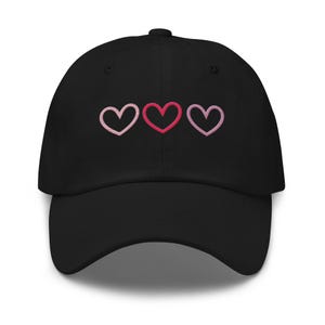 May include: Black baseball cap with three embroidered hearts in shades of pink. The hearts are arranged horizontally across the front of the cap. The cap has a curved brim and an adjustable strap.