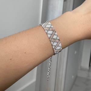 May include: A beaded bracelet with a geometric pattern of white, tan, and silver beads. The bracelet is on a person's wrist. The bracelet has a silver chain for adjusting the size.