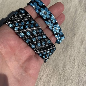 May include: Two beaded bracelets. One is black with a blue floral pattern, and the other is blue with a floral pattern. The bracelets are made of small beads and are held in a hand.