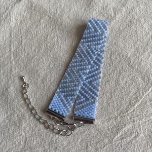 Könnte beinhalten: Ein hellblaues gewebtes Perlenarmband mit geometrischem Zickzackmuster. Das Armband ist mit silberfarbenen Metallendkappen und einer silberfarbenen Metallverlängerungskette mit Karabinerverschluss zur individuellen Größenanpassung versehen.