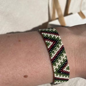 May include: A beaded bracelet with a geometric pattern of white, green, and burgundy beads. The bracelet is worn on a wrist and has a clasp closure. The design features diagonal stripes.