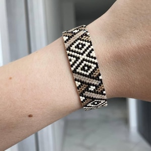 May include: A beaded bracelet with a geometric pattern in black, white, and tan. The bracelet is worn on a wrist and features a woven design. The bracelet is approximately 1.5 inches wide.