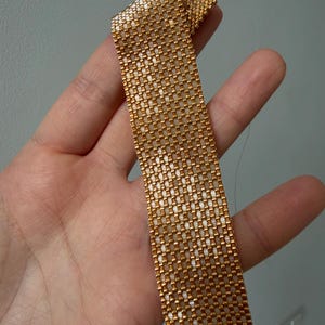 May include: A flat, rectangular strip of woven metallic gold beads. The beadwork features a consistent grid pattern with a reflective, shiny finish. The strip is flexible and appears to be a segment of a larger piece of jewelry or accessory.