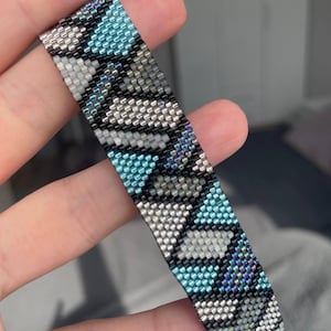May include: A beaded bracelet with a geometric pattern in shades of blue, gray, black, and iridescent beads. The bracelet is held up, showcasing the intricate design and craftsmanship. The beads are small and tightly woven together.