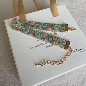 May include: A beaded choker necklace with a geometric pattern in shades of blue, tan, and pink, with gold-colored clasps and chain. The necklace rests on a white gift bag with a gold ribbon.