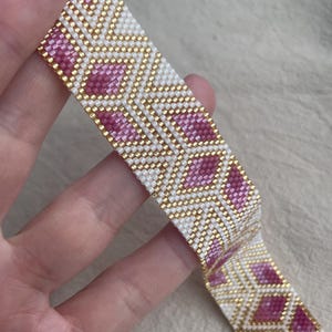 May include: A woven bead bracelet featuring a geometric pattern of white, metallic gold, and magenta diamond shapes. The intricate design is created with small, uniform beads arranged in a repeating, symmetrical structure across the entire band.