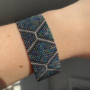 May include: A wide beaded bracelet with a geometric pattern. The bracelet features a mix of dark gray, iridescent blue, and green beads. The design creates a textured, eye-catching accessory.