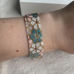 May include: A beaded bracelet featuring a geometric design in pink, teal, white, and gold beads. The bracelet is worn on a wrist. The beads are small and tightly woven, creating a textured surface. The bracelet is likely handmade.