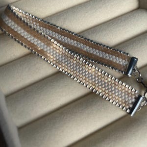 Puede incluir: Una pulsera de cuentas tejidas con rayas horizontales de cuentas color beige, blanco y plata iridiscente. La pulsera está rematada con terminales de metal plateado y un cierre a juego, creando un aspecto estructurado y uniforme.
