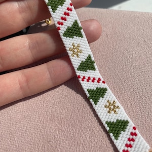 Pode incluir: Uma pulseira de contas tecidas com fundo branco e um padrão repetitivo de árvores de Natal verdes, formas de estrelas douradas e listras diagonais vermelhas. A pulseira é finalizada com terminais de metal dourado e um fecho.