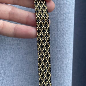 May include: A narrow, rectangular bracelet crafted from small, uniform beads arranged in a repeating geometric pattern. The design features alternating vertical columns of metallic gold and matte black beads, creating a structured, woven appearance.