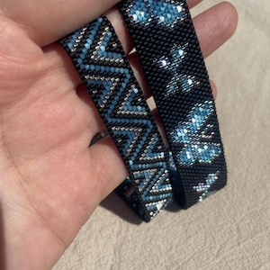 May include: Two beaded bracelets made with small square beads. One bracelet features a repeating blue and silver chevron pattern on a black background. The other bracelet displays a series of blue butterfly shapes against a solid black background.