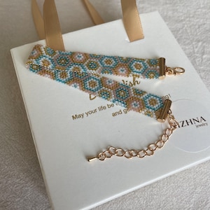 May include: A beaded choker necklace with a geometric pattern in shades of blue, tan, and pink, with gold-colored metal clasps and chain. The necklace rests on a white gift box with gold ribbon handles. Text on the box reads "May your life be..." and "ZHNA jewelry".