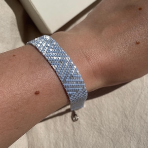 May include: A light blue and white beaded bracelet with a silver-colored clasp. The bracelet is made of small, square beads arranged in a woven pattern. The bracelet is shown on a person's wrist.