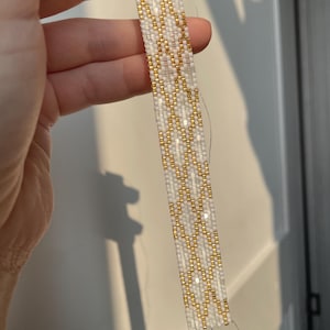 May include: A long, narrow beaded bracelet or strip featuring a repeating geometric diamond pattern. The design is crafted from small, iridescent white beads and metallic gold beads, creating a structured, woven appearance with visible thread.