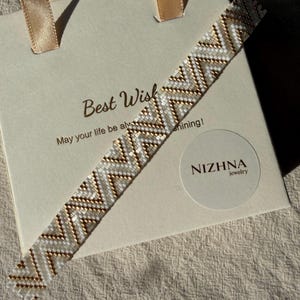 May include: A beaded bracelet featuring a geometric chevron pattern in alternating white and metallic gold beads. The bracelet is displayed diagonally across a cream-coloured gift bag with the text Best Wish May your life be always shining and NIZHNA jewelry.