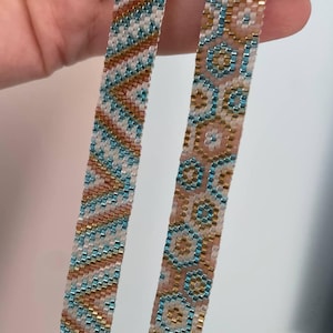May include: Two beaded bracelets with intricate geometric patterns. One features a chevron design in teal, gold, and white. The other displays a floral-like pattern in teal, gold, and pink. The bracelets are held against a white background.