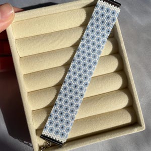 May include: A beaded bracelet with a floral pattern in light blue, white, and black. The bracelet has a silver clasp and is displayed on a beige jewelry box. The bracelet is approximately 1 inch wide.