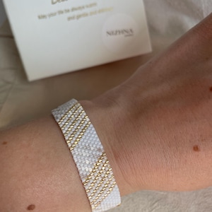 Op de afbeelding: Wit en goud kralenarmband met een geometrisch patroon. De armband zit om een pols. Op de achtergrond is een witte doos met de tekst "Best Wish" te zien.