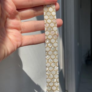 Puede incluir: Una pulsera de cuentas con un patrón de panal repetitivo creado con pequeñas cuentas de oro metálico y cuentas blancas translúcidas. La banda rectangular está terminada con un cierre de metal dorado y una cadena extensora corta.
